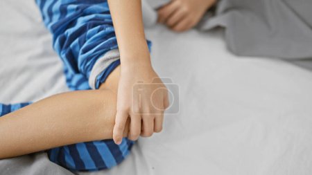 Téléchargez les photos : Adorables mains d'enfant touchant genou blessé dans la douleur, souffrant couché dans le lit dans une chambre à coucher. réveillé tôt le matin, le garçon bouleversé en pyjama montre son inquiétude à cause d'un problème d'articulation du genou. - en image libre de droit