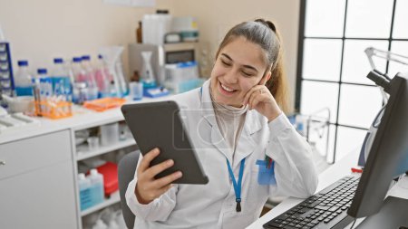 Mujer joven sonriente con bata blanca de laboratorio que usa tabletas en un entorno de laboratorio.