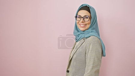 Téléchargez les photos : Une femme d'âge moyen souriante portant un hijab et des lunettes isolées contre un mur rose. - en image libre de droit