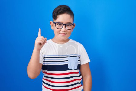 Foto de Joven niño hispano de pie sobre fondo azul mostrando y señalando con el dedo número uno mientras sonríe confiado y feliz. - Imagen libre de derechos