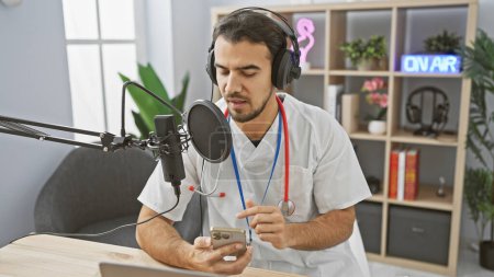 Hombre hispano guapo en podcasting de uniforme médico con micrófono y teléfono inteligente en estudio de radio.