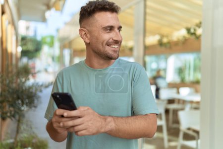 Téléchargez les photos : Jeune homme hispanique souriant confiant en utilisant smartphone à la terrasse du café - en image libre de droit