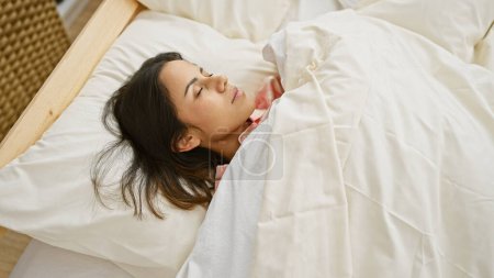 Téléchargez les photos : Une jeune femme hispanique sereine dort dans un lit blanc confortable, mettant en valeur la tranquillité et le confort dans un cadre de chambre à coucher accueillant. - en image libre de droit