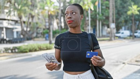 Téléchargez les photos : Femme afro-américaine sérieuse tenant une carte de crédit et des dollars dans la rue de la ville - en image libre de droit
