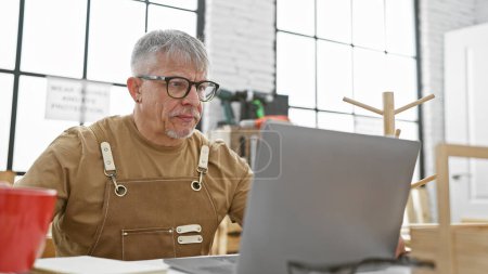 Téléchargez les photos : Un homme mûr aux cheveux gris s'assoit dans un atelier, se concentrant intensément sur un écran d'ordinateur portable entouré d'outils de menuiserie. - en image libre de droit