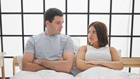 Téléchargez les photos : Beau couple assis sur le lit avec les bras croisés geste regardant bouleversé à la chambre - en image libre de droit