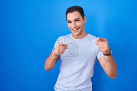 Photo for Young hispanic man standing over blue background pointing fingers to camera with happy and funny face. good energy and vibes. - Royalty Free Image