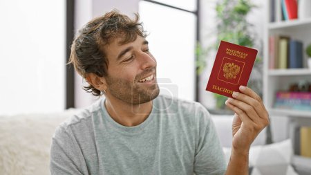 Téléchargez les photos : Jeune homme joyeux visite son salon, tenant joyeusement haut un passeport russe, détendu assis sur le canapé - en image libre de droit