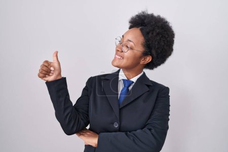 Photo for Beautiful african woman with curly hair wearing business jacket and glasses looking proud, smiling doing thumbs up gesture to the side - Royalty Free Image