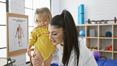 Téléchargez les photos : Une pédiatre examine le genou d'une petite fille dans une salle de physiothérapie éclairée avec un équipement de réadaptation. - en image libre de droit