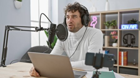 Photo for Bearded hispanic man recording podcast in modern studio with laptop and microphone. - Royalty Free Image