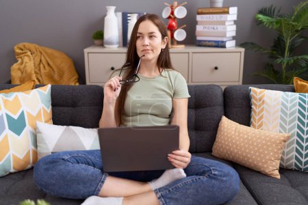 Téléchargez les photos : Jeune belle femme utilisant un ordinateur portable assis sur le canapé avec l'expression du doute à la maison - en image libre de droit