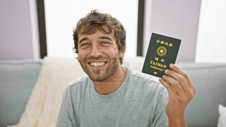 Photo for Beaming young man, confidently enjoying his staycation on the sofa, holding a taiwan passport, in a cozy home setting - Royalty Free Image