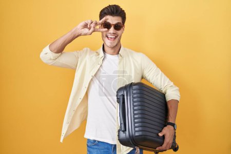 Foto de Joven hombre hispano sosteniendo maleta yendo de vacaciones de verano haciendo símbolo de paz con los dedos en la cara, sonriendo alegre mostrando la victoria - Imagen libre de derechos