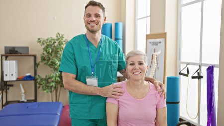 Photo for Physiotherapist, a man in green scrubs, standing with woman patient in pink shirt in a bright physiotherapy clinic room. - Royalty Free Image