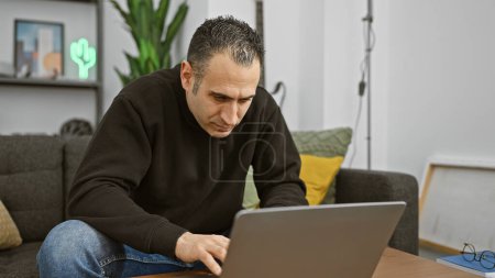 Téléchargez les photos : Homme hispanique concentré utilisant un ordinateur portable dans un cadre de salon moderne - en image libre de droit