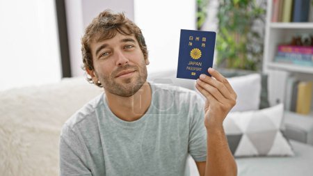 Photo for Confident young man, joy radiating from his smile, sitting casual on his sofa, holding passport, dreaming of his next adventure in japan right from home - Royalty Free Image
