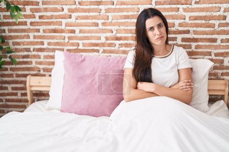 Foto de Young beautiful hispanic woman sitting on bed with unhappy expression and arms crossed gesture at bedroom - Imagen libre de derechos