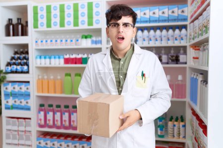 Téléchargez les photos : Jeune homme non binaire travaillant à la pharmacie drugstore tenant boîte effrayé et étonné avec la bouche ouverte pour la surprise, visage incrédule - en image libre de droit