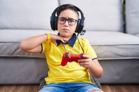 Téléchargez les photos : Jeune enfant hispanique jouant contrôleur de tenue de jeu vidéo portant des écouteurs tranchant la gorge avec la main comme couteau, menacer d'agression avec une violence furieuse - en image libre de droit