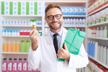 Téléchargez les photos : Homme d'âge moyen avec barbe travaillant à la pharmacie drugstore tenant des pilules souriant avec un sourire heureux et cool sur le visage. montrant les dents. - en image libre de droit