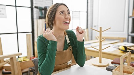 Foto de Una mujer alegre en un suéter verde y delantal marrón sonríe en un taller de carpintería bien iluminado con varias herramientas de carpintería. - Imagen libre de derechos