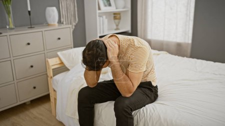 Photo for Upset young hispanic man sitting on bed in bedroom, holding his head in distress. - Royalty Free Image