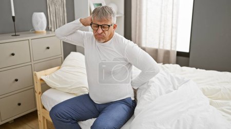 Téléchargez les photos : Un homme aux cheveux gris endolori, assis sur un lit dans une chambre bien éclairée - en image libre de droit