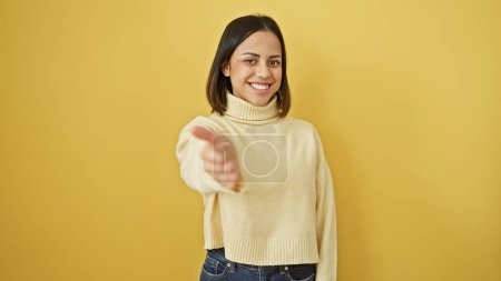 Photo for A smiling young hispanic woman extends her hand in greeting against an isolated yellow background. - Royalty Free Image