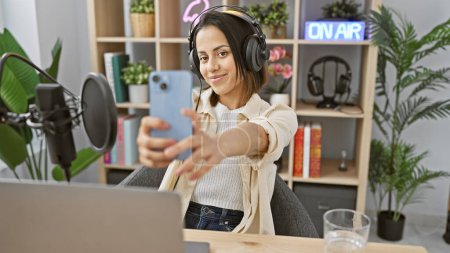 Una joven hispana se toma una selfie en un estudio de radio con auriculares y un letrero 'en el aire' en el fondo.