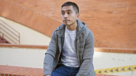 Un joven contemplativo hispano se sienta en las escaleras urbanas, exudando un ambiente casual y guapo en un ambiente de ciudad.