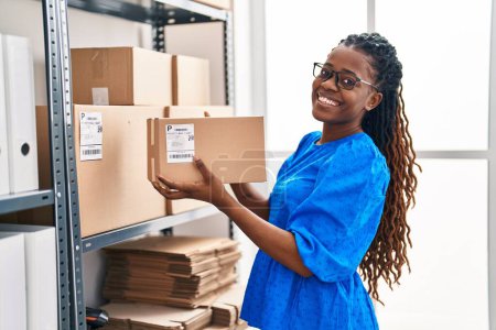 Téléchargez les photos : Femme afro-américaine commerce électronique travailleur d'entreprise tenant paquet d'étagères au bureau - en image libre de droit