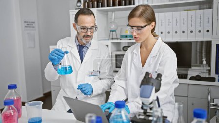 Photo for Two jubilant scientists working together in a lab, conversing and using a laptop while holding a test tube for sample analysis in their indoor science research workplace. - Royalty Free Image