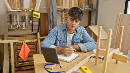 Photo for Hardworking young hispanic man masterfully navigating carpentry business, deftly taking notes on touchpad amid timber and furniture in busy workshop - Royalty Free Image