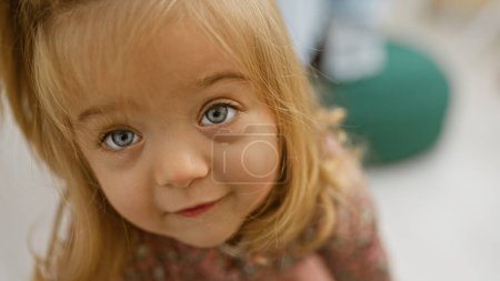 Photo for Close-up of a curious little girl with blonde hair and blue eyes indoors, evoking a sense of innocence and childhood. - Royalty Free Image