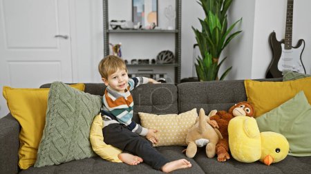 Téléchargez les photos : Petit garçon blond souriant sur un canapé gris avec des oreillers colorés et des animaux en peluche dans un salon moderne. - en image libre de droit