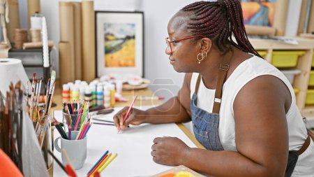 Téléchargez les photos : Femme afro-américaine focalisée, artiste captivante, dessinant avec concentration sur son carnet dans un studio d'art d'intérieur. - en image libre de droit