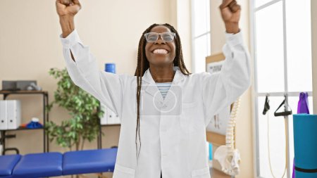 Photo for African american woman with braids wearing lab coat celebrates success in clinic room - Royalty Free Image