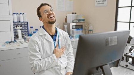 Photo for Laughing hispanic man in lab coat sitting at computer in bright laboratory setting. - Royalty Free Image