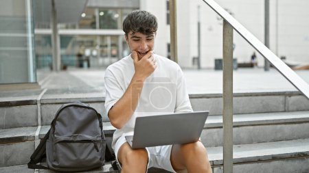 Photo for Cool, confident young hispanic male student, lifestyle portrait of a handsome teenager smiling, sitting on university stairs using laptop in sunlit city backdrop, indulging in online education. - Royalty Free Image