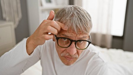 Photo for A thoughtful senior man with glasses touching his forehead wrinkles in a white bedroom. - Royalty Free Image