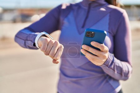 Téléchargez les photos : Jeune belle femme hispanique portant des vêtements de sport en utilisant smartphone regarder chronomètre à la rue - en image libre de droit