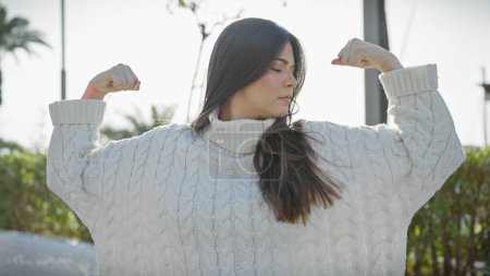 Photo for Hispanic woman flexing muscles confidently in a park with greenery wearing a white sweater. - Royalty Free Image