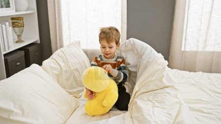 Foto de Un niño rubio juega con un juguete de peluche amarillo en una cama blanca en un acogedor dormitorio soleado - Imagen libre de derechos