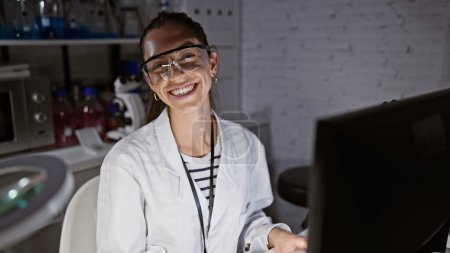 Téléchargez les photos : Souriante, confiante jeune femme hispanique scientifique, belle dans ses lunettes, appréciant son travail sur l'ordinateur au laboratoire - en image libre de droit