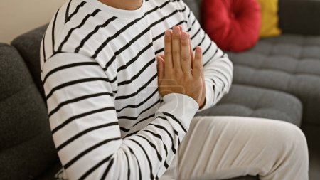 Téléchargez les photos : La prière intérieure émotive de l'homme hispanique, assis sur le canapé du salon à la maison, se détendre dans la méditation et trouver l'équilibre dans la foi - en image libre de droit