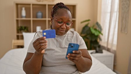 Téléchargez les photos : Femme afro-américaine souriante assise confortablement dans sa chambre ; profiter des achats en ligne le matin sur le smartphone, en utilisant une carte de crédit dans sa chambre intérieure confortable - en image libre de droit