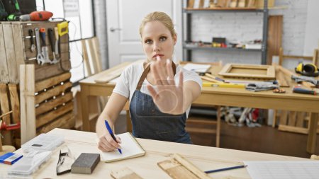 Foto de Una joven en un delantal de mezclilla hace gestos para detenerse mientras planea la carpintería en un estudio de carpintería. - Imagen libre de derechos
