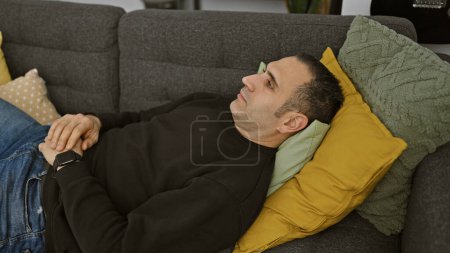 Téléchargez les photos : Homme hispanique détendu couché sur un canapé dans un intérieur confortable avec des oreillers décoratifs. - en image libre de droit