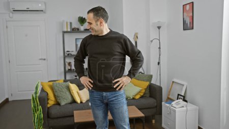 Selbstbewusster hispanischer Mann, der in einem gut ausgestatteten modernen Wohnzimmer mit den Händen auf den Hüften steht und lächelnd wegschaut.